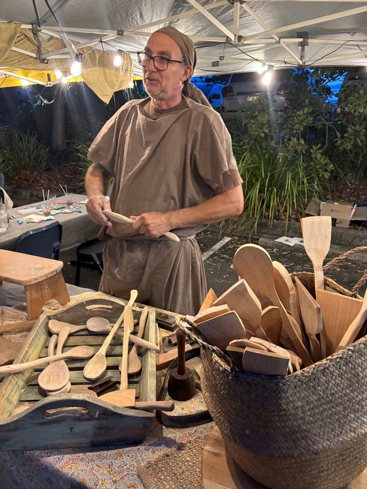 A carpenter in costume at his stall, surrounded by hand-carved wooden spoons and tools