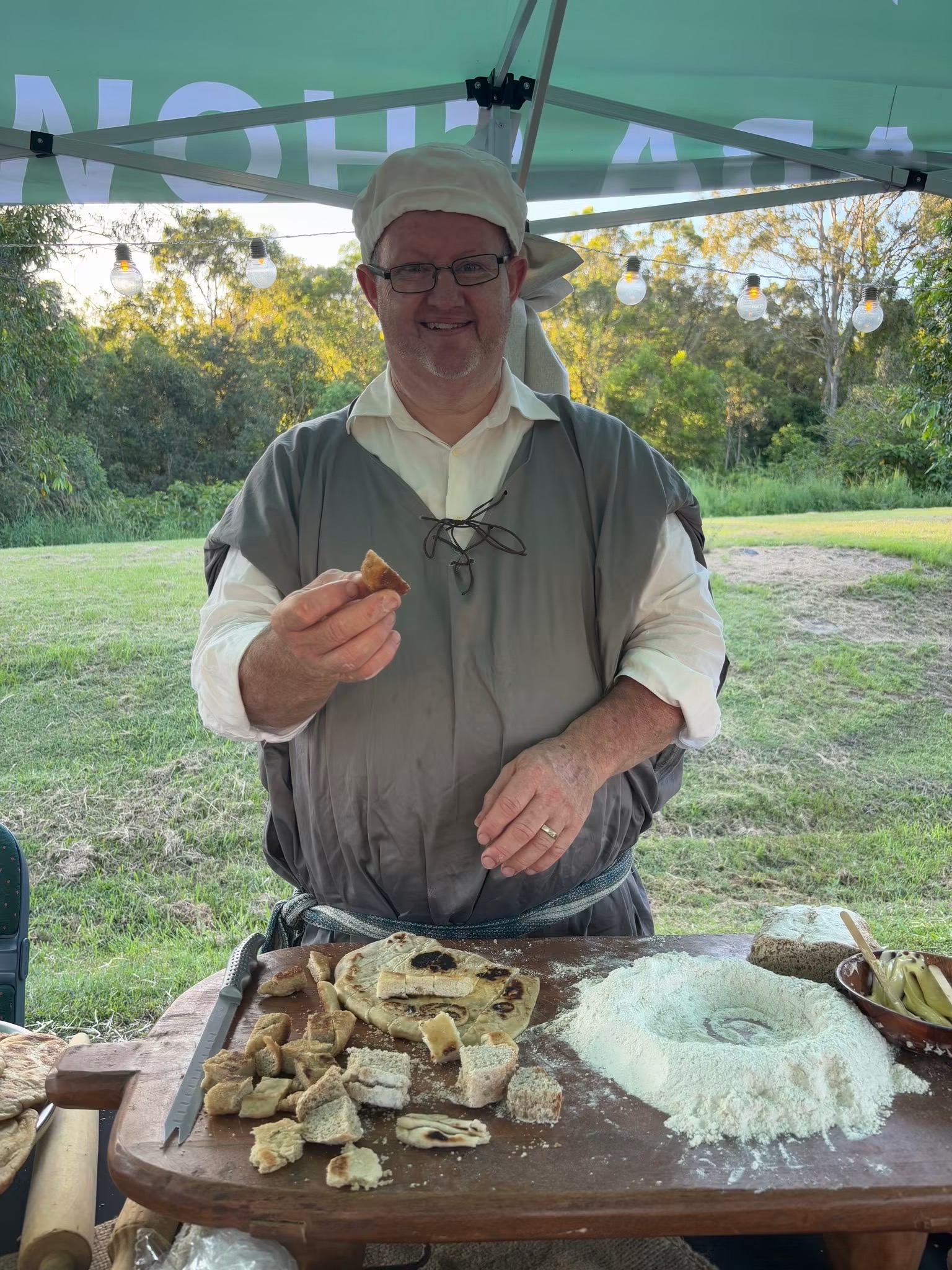 A baker in costume holding a piece of flatbread beside an outdoor oven