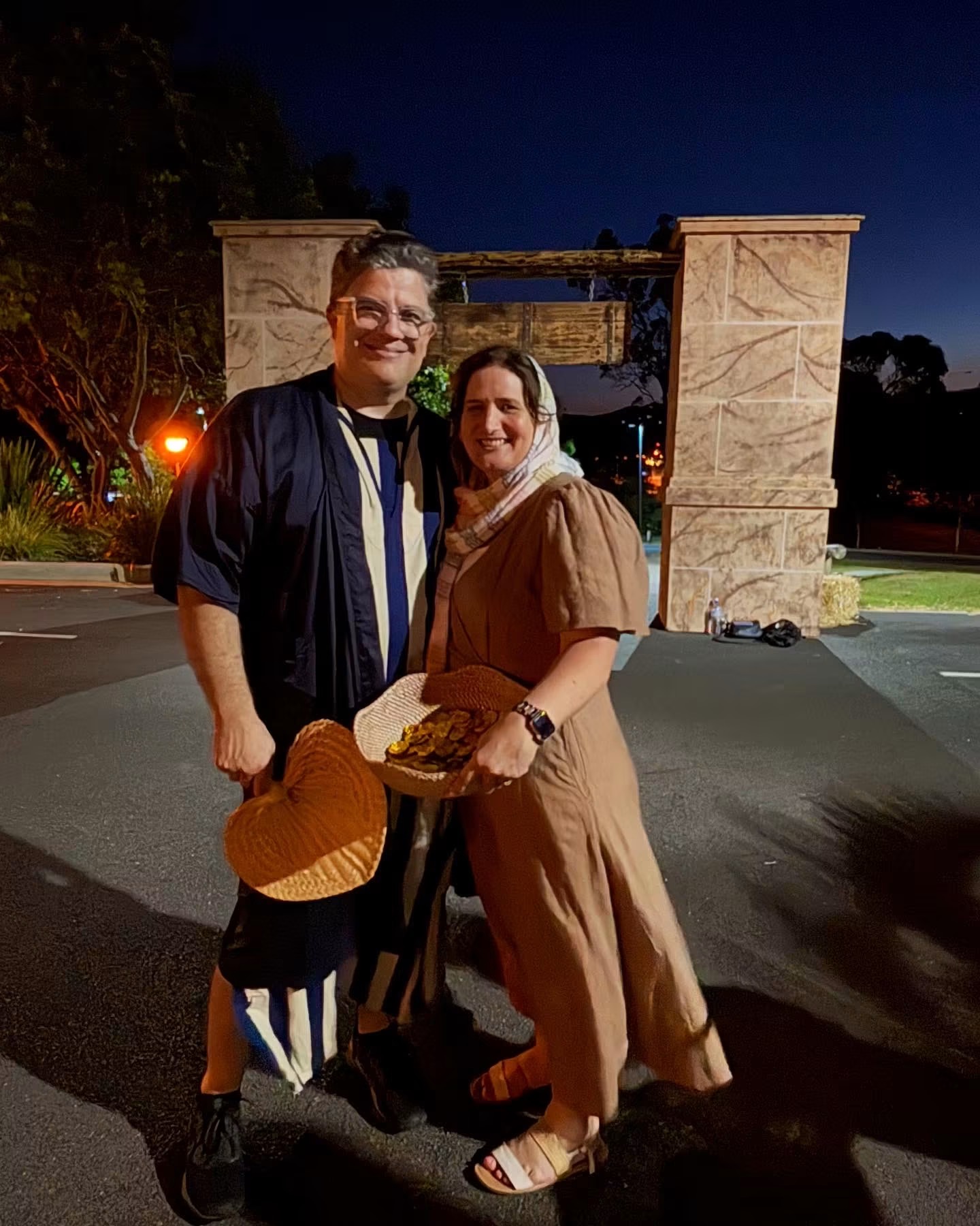 Two villagers in costume carrying woven baskets at night beside the stone archway into the town