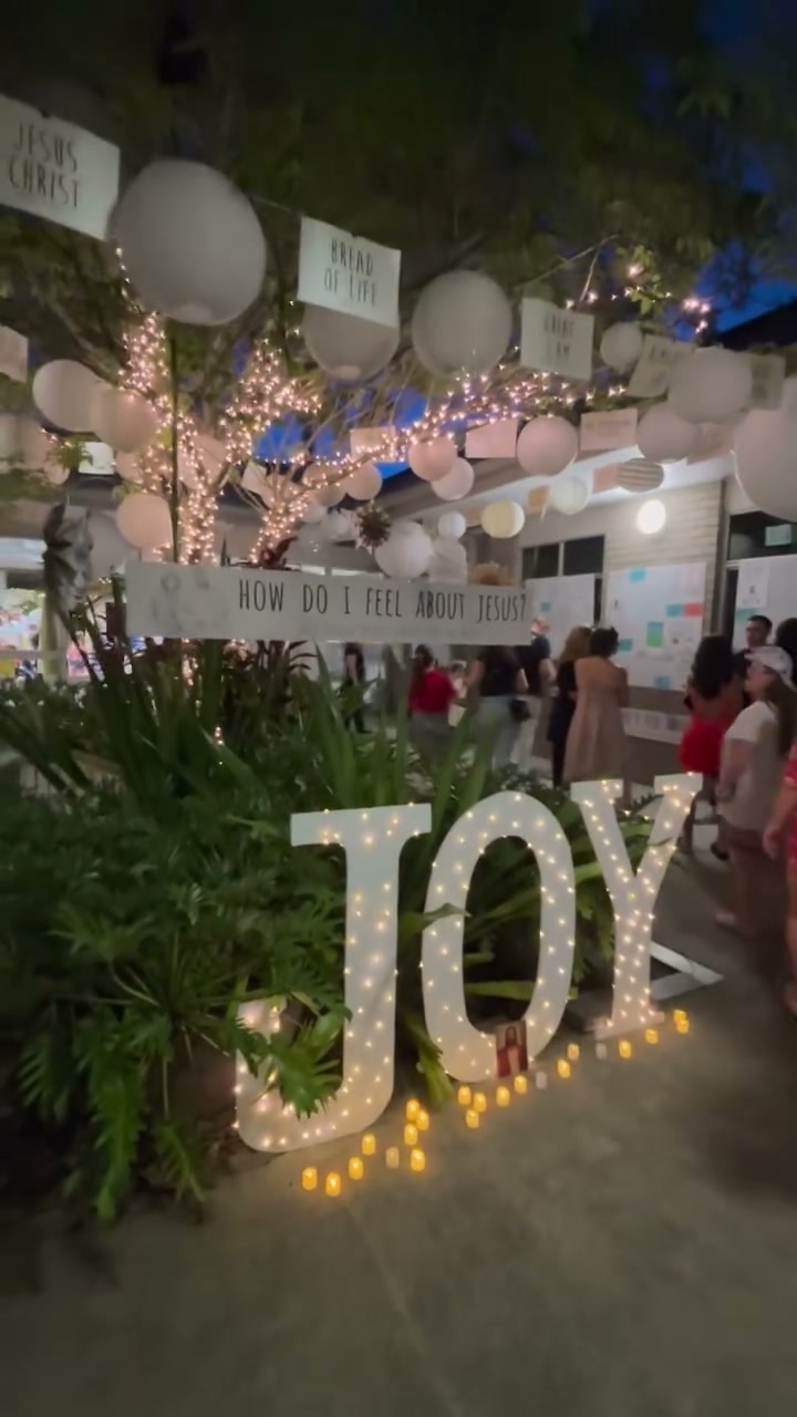 Illuminated JOY letters among candles and potted palms, under paper lanterns and fairy lights
