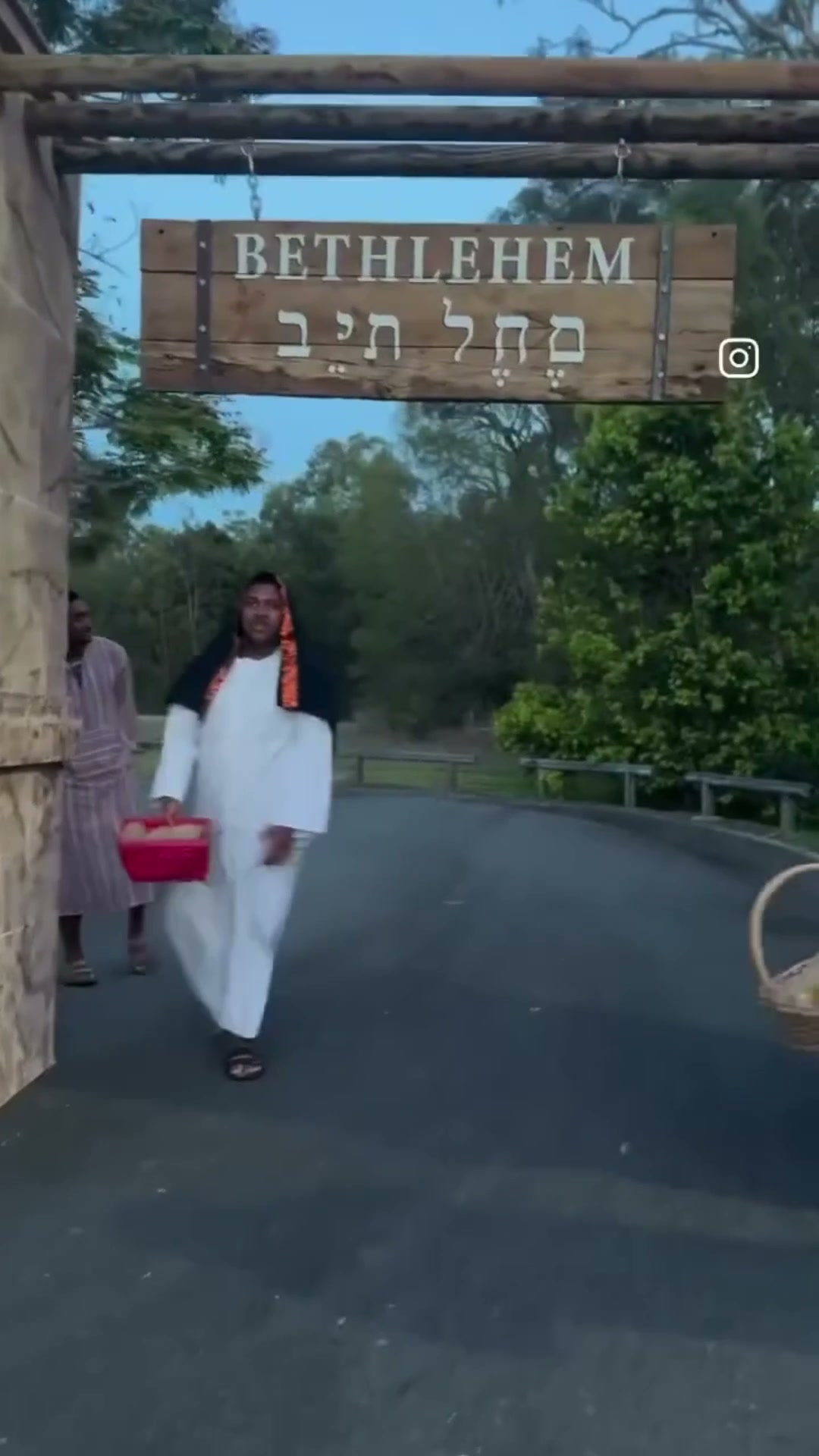 Two villagers in costume carrying baskets, walking out under the timber archway whose hanging sign reads Bethlehem in English and Hebrew